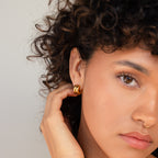 A woman with curly hair touches her ear, showing off the Chunky Double Hoops—a bold statement accessory—with a neutral expression.