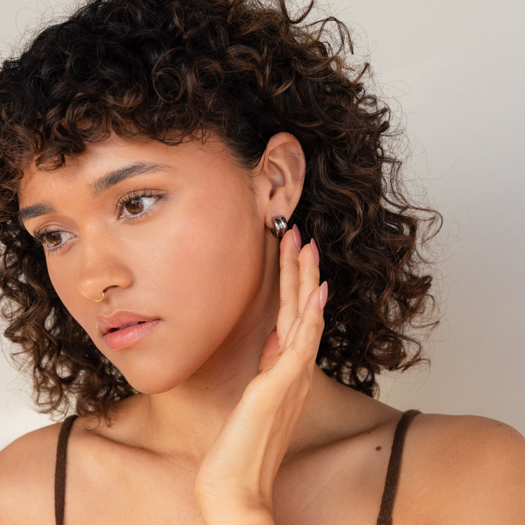 A woman with curly hair wears Chunky Double Hoops, touches her ear, and gazes thoughtfully to the side—showcasing how these earrings make a striking minimalist statement.
