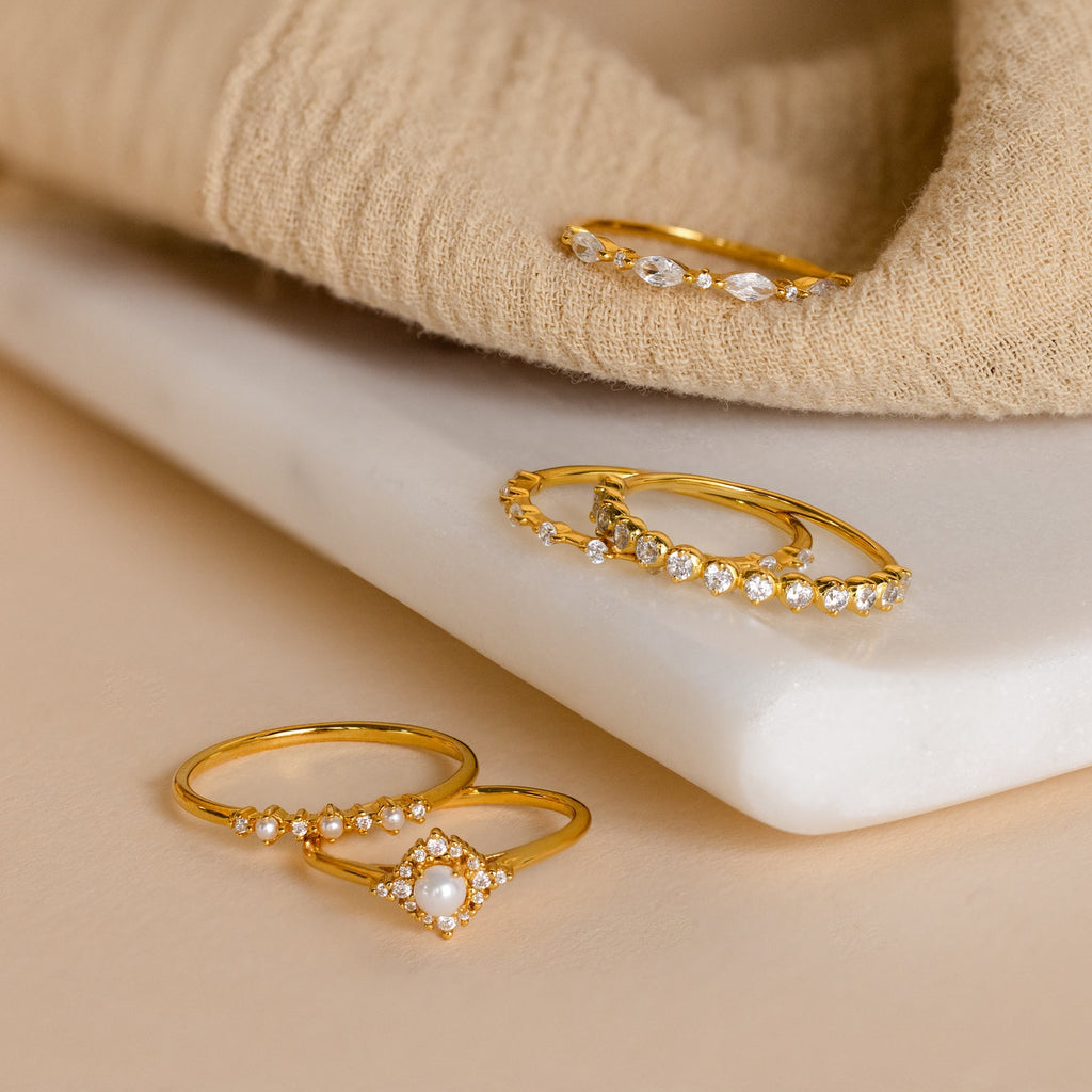 Four elegant Pearl Diamond Stacking Rings, featuring pearls and small gemstones, are displayed on a white stone and beige fabric background.