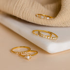 Four elegant Pearl Diamond Stacking Rings, featuring pearls and small gemstones, are displayed on a white stone and beige fabric background.
