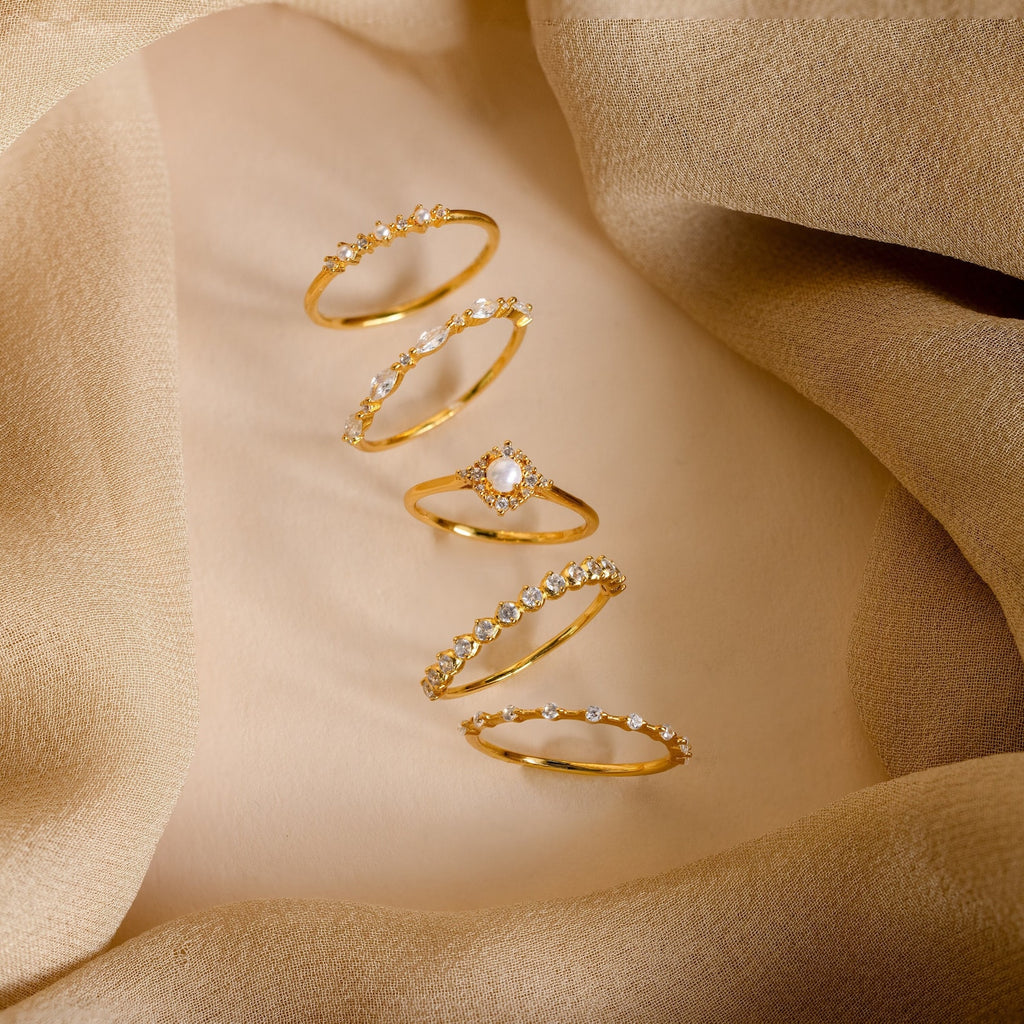 Five gold rings with gemstones, including Pearl Diamond Stacking Rings and a Crystal Stacking Ring Set, are displayed on a beige fabric background.