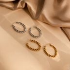 Two pairs of Bold Twisted Hoops, one silver and one gold, featuring a sleek twist design, elegantly displayed on beige satin fabric.
