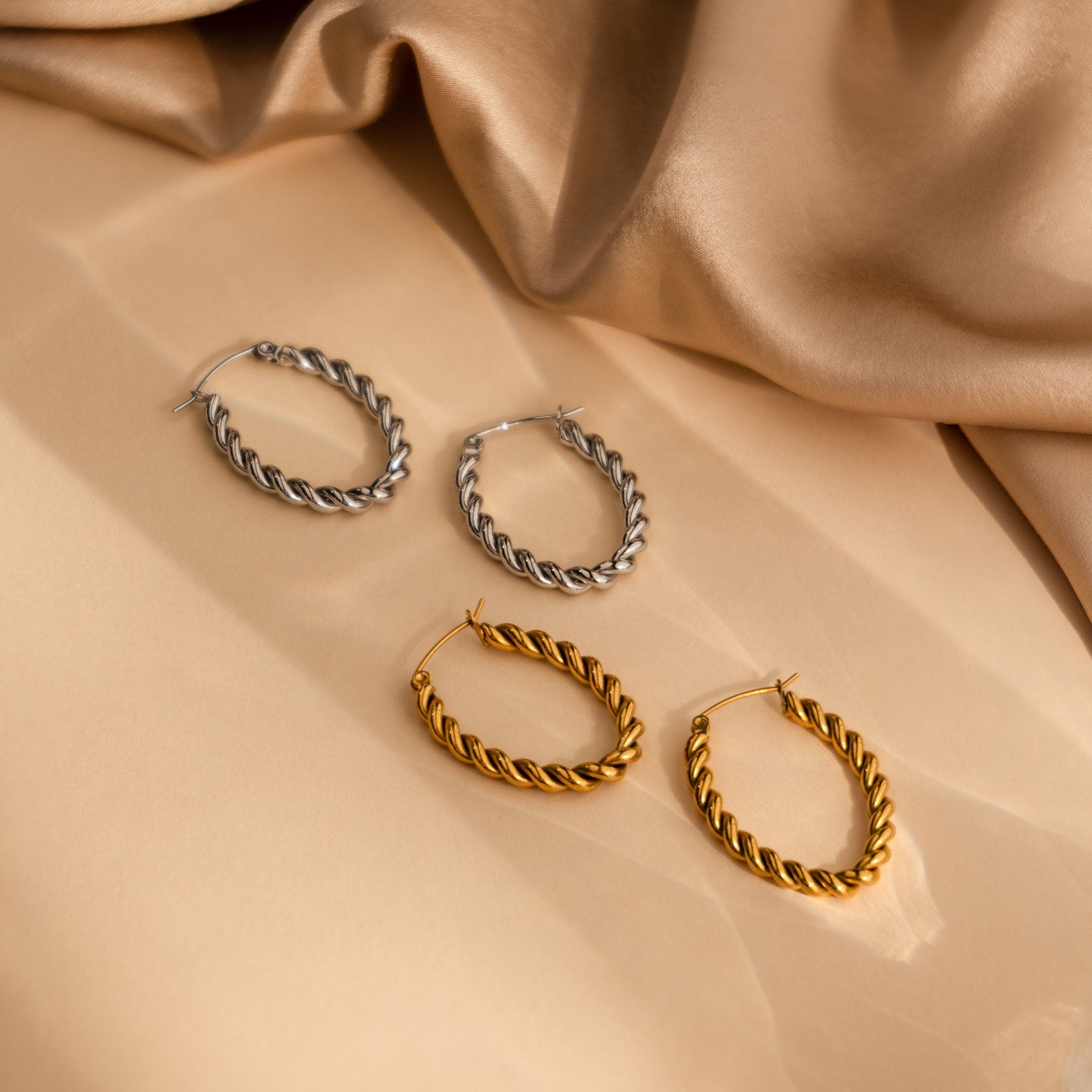 Two pairs of Bold Twisted Hoops, one silver and one gold, featuring a sleek twist design, elegantly displayed on beige satin fabric.