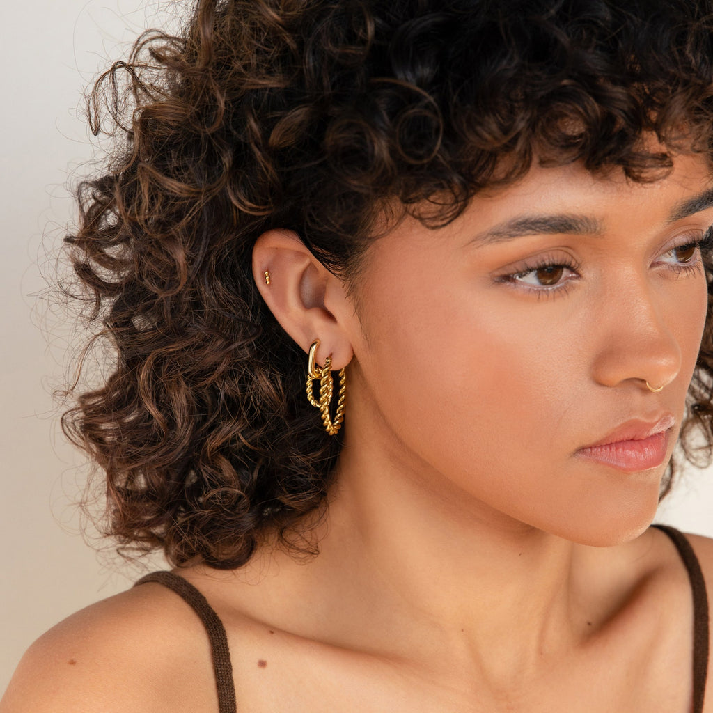 A woman with short curly hair and a nose ring looks to the side, wearing Bold Twisted Hoops.