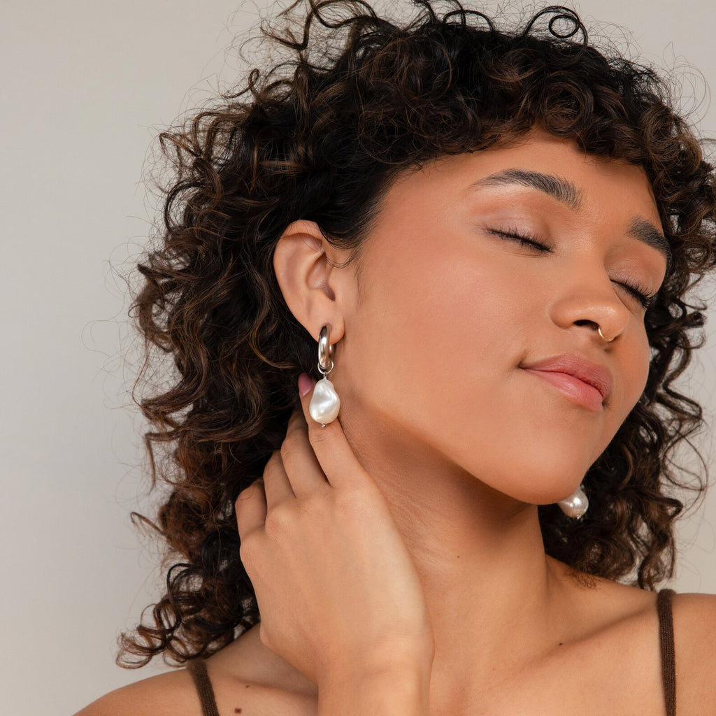 A woman with curly hair wears elegant Baroque Pearl Drop Hoops, adding a touch of sophistication to her look.