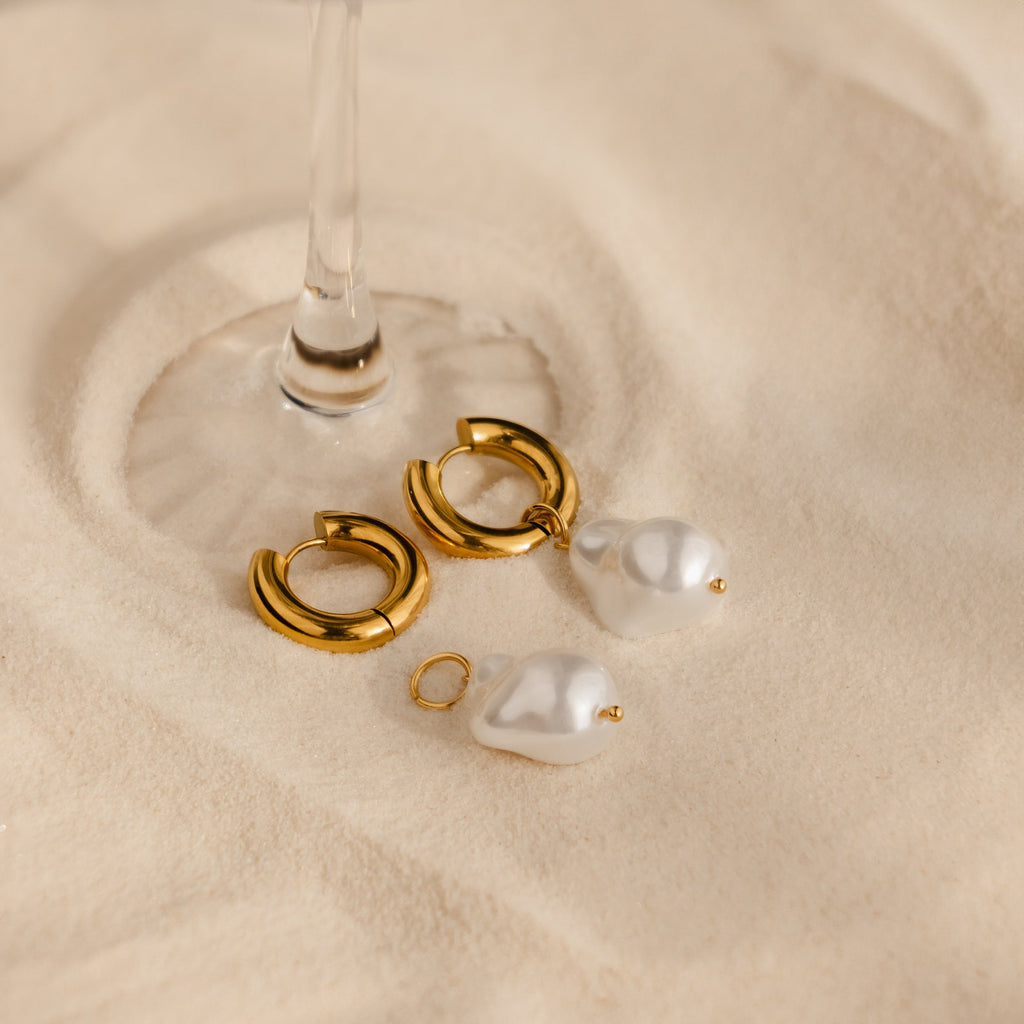 Baroque Pearl Drop Hoops—gold hoops with detachable baroque pearls—rest on a beige surface under a glass stem, making striking statement earrings for your wedding jewelry collection.