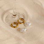 Baroque Pearl Drop Hoops—gold hoops with detachable baroque pearls—rest on a beige surface under a glass stem, making striking statement earrings for your wedding jewelry collection.