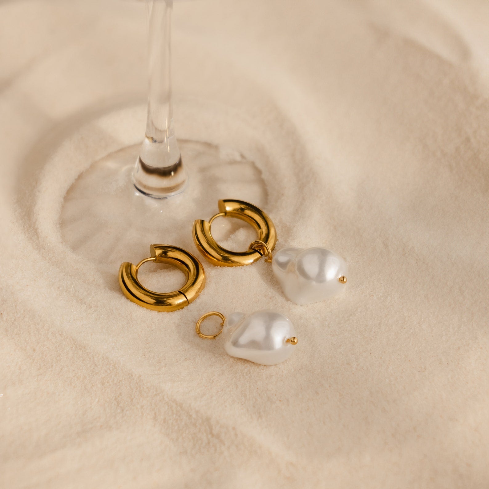 Baroque Pearl Drop Hoops—gold hoops with detachable baroque pearls—rest on a beige surface under a glass stem, making striking statement earrings for your wedding jewelry collection.