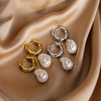 Displayed on beige satin, two pairs of Baroque Pearl Drop Hoops—one gold, one silver—feature hoop designs with dangling pearls. These statement earrings are perfect as elegant wedding jewelry for any occasion.