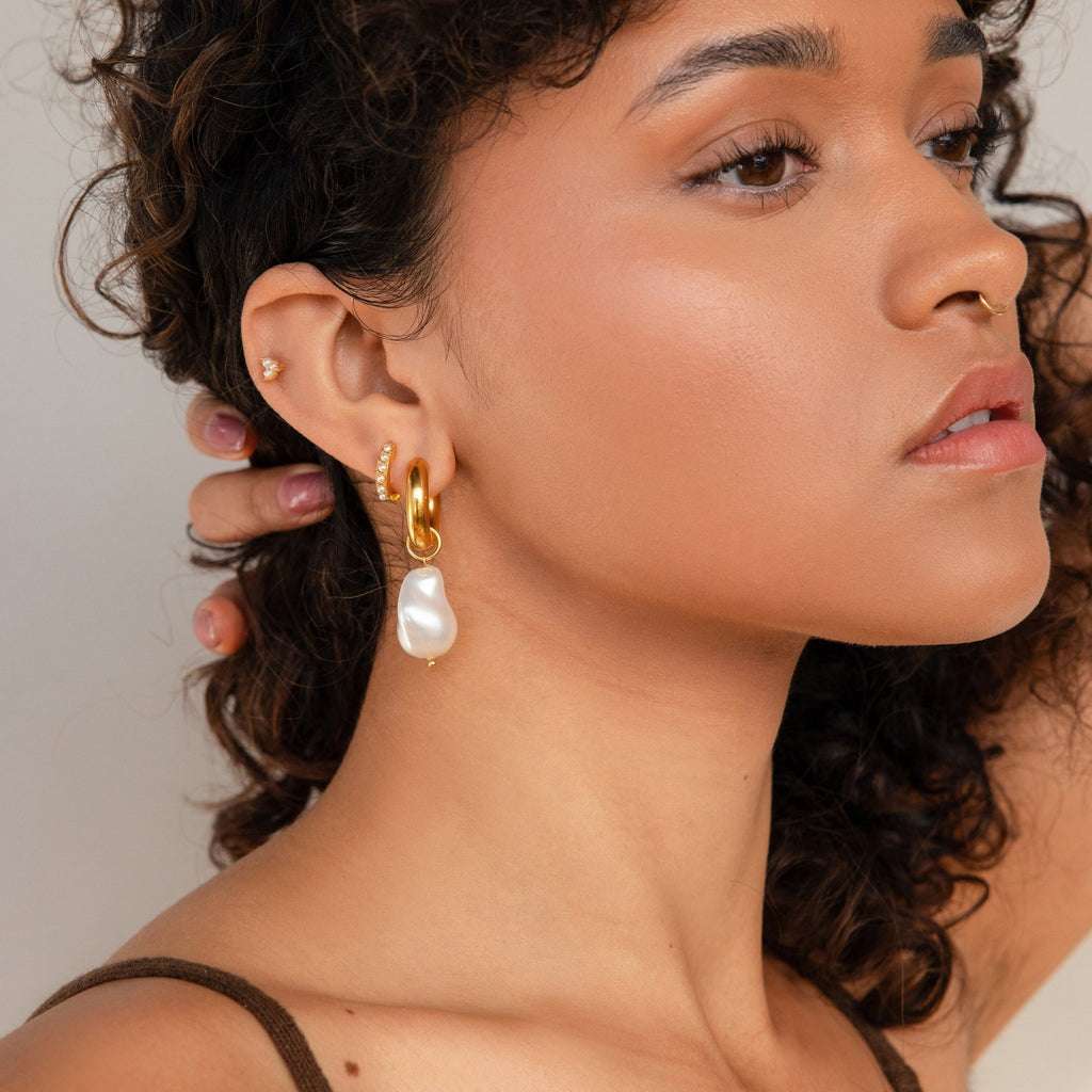 A woman with curly hair and glowing skin wears Baroque Pearl Drop Hoops and a nose ring, her small ear piercings accentuating these statement earrings—ideal for elevating both everyday looks and elegant wedding styles.