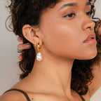 A woman with curly hair and glowing skin wears Baroque Pearl Drop Hoops and a nose ring, her small ear piercings accentuating these statement earrings—ideal for elevating both everyday looks and elegant wedding styles.