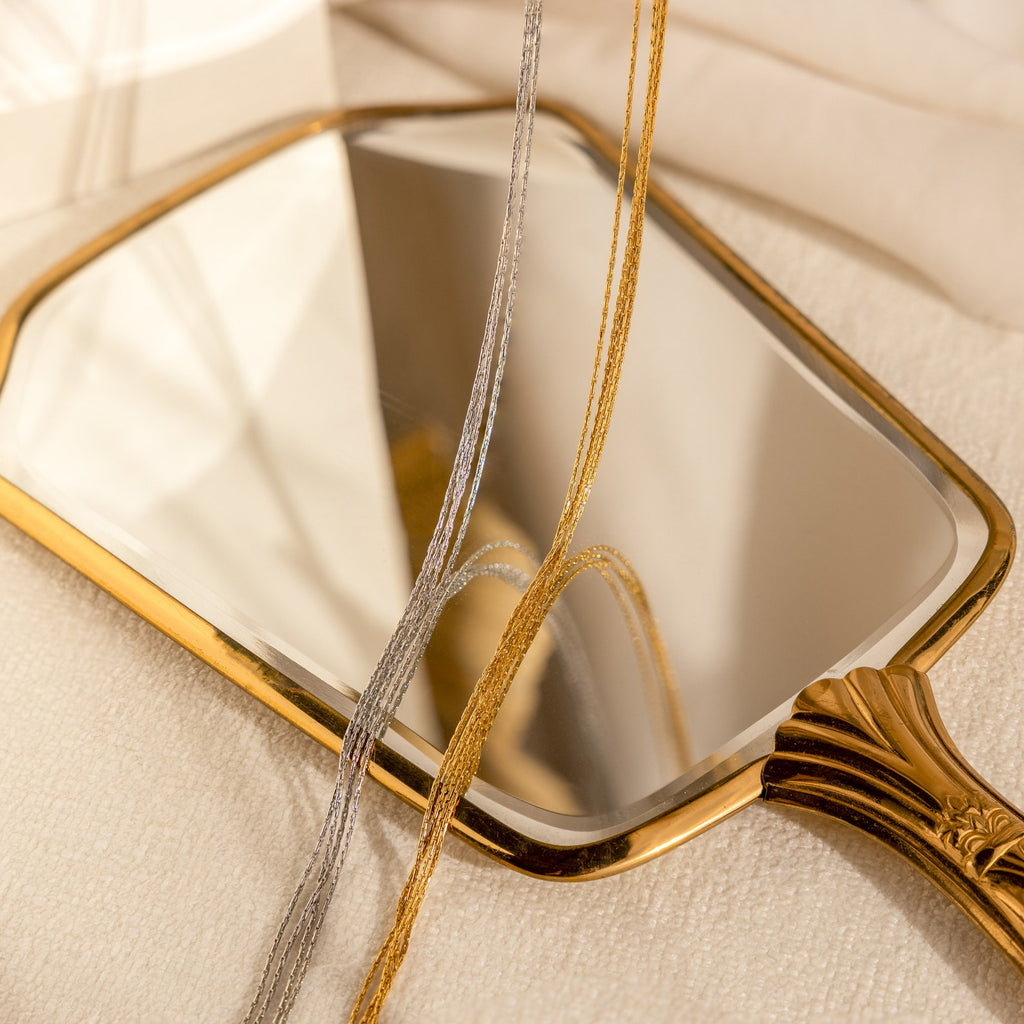 A Multi Strand Chain Necklace rests on a soft surface, draped over a gold-handled hand mirror that reflects light, paired with layered silver and gold necklaces for a minimalistic look.