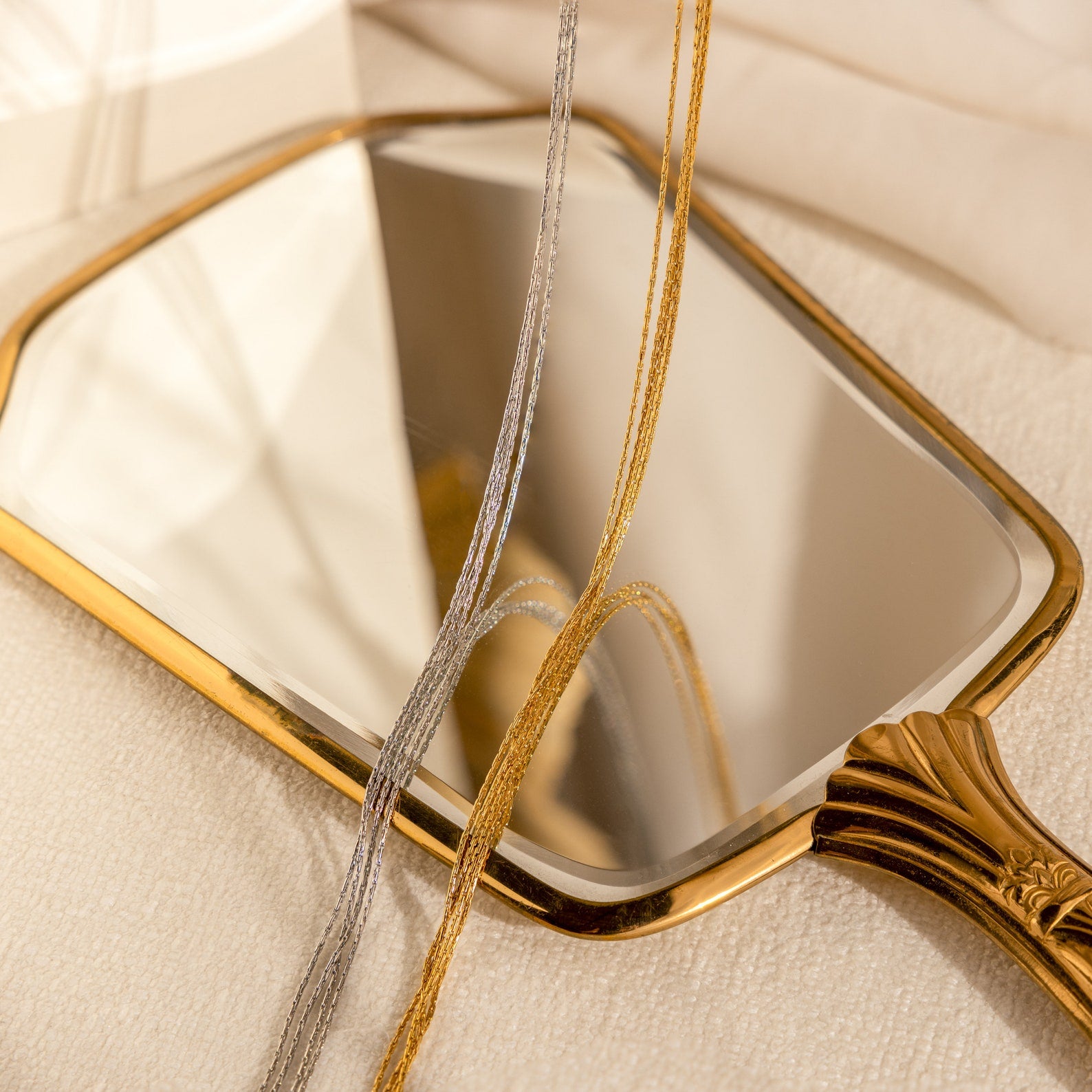 A Multi Strand Chain Necklace rests on a soft surface, draped over a gold-handled hand mirror that reflects light, paired with layered silver and gold necklaces for a minimalistic look.