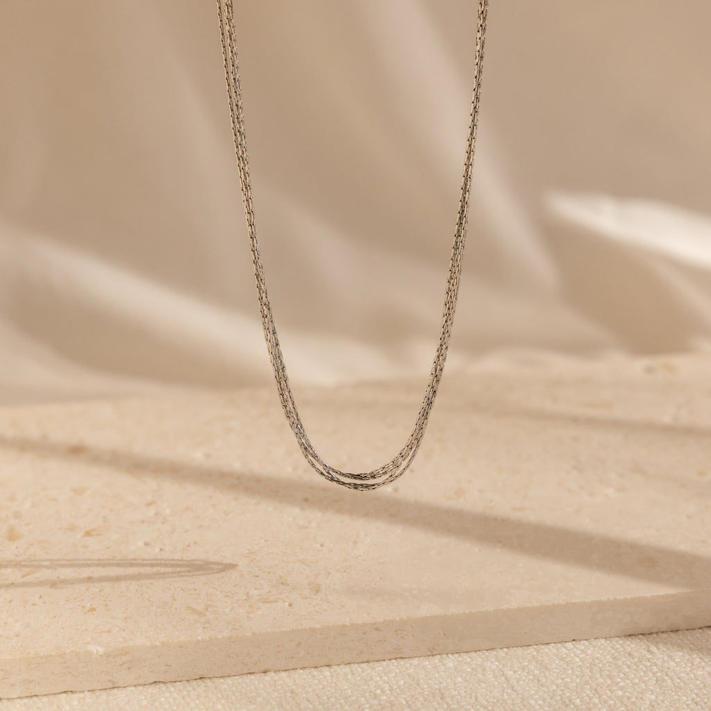 The Multi Strand Chain Necklace is a delicate, minimalistic jewelry piece that hangs gracefully against a neutral, textured background.