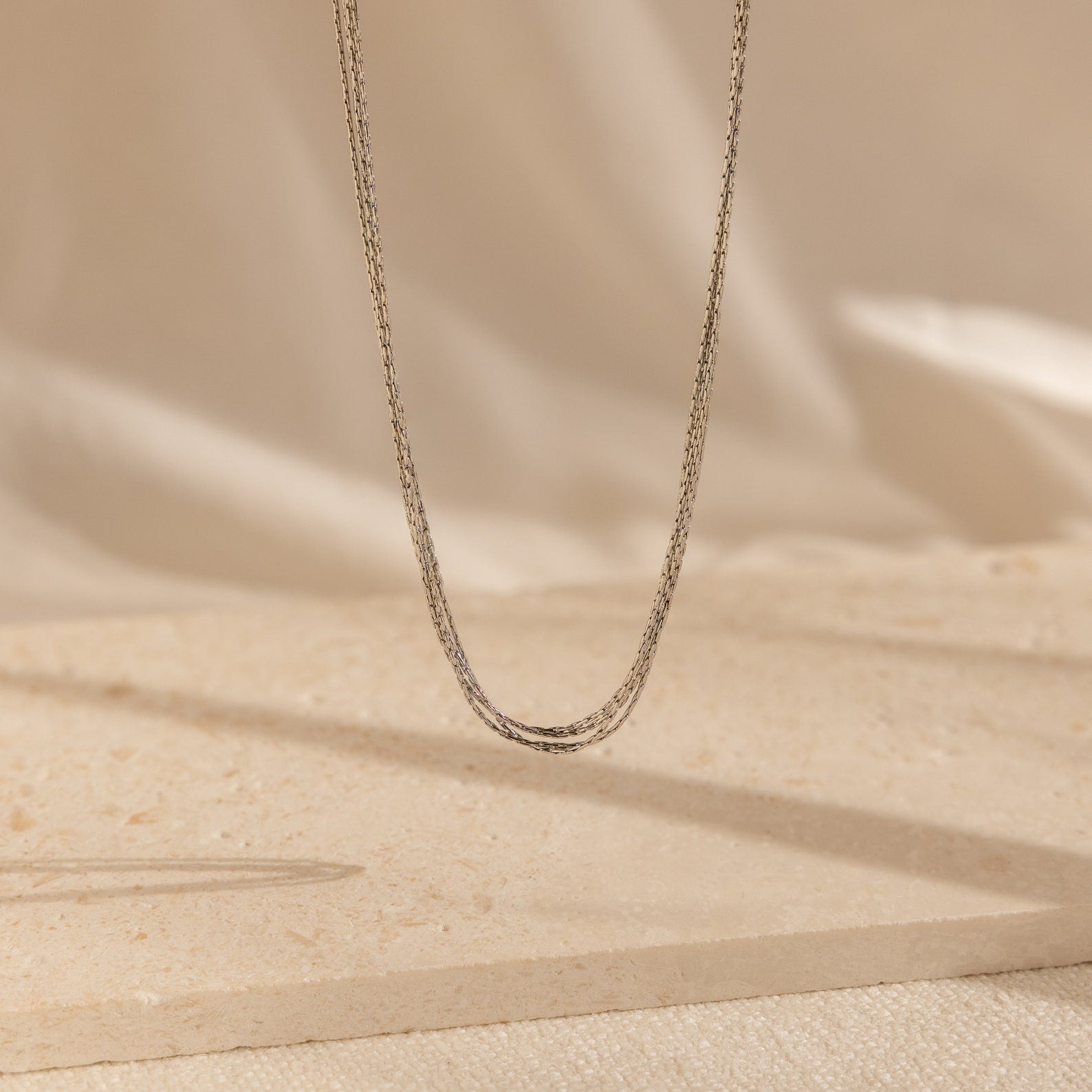 The Multi Strand Chain Necklace is a delicate, minimalistic jewelry piece that hangs gracefully against a neutral, textured background.