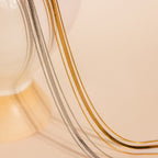 The Triple Layered Chain Necklace in gold and silver is elegantly draped over the edge of a cream-colored vase, set against a beige backdrop.