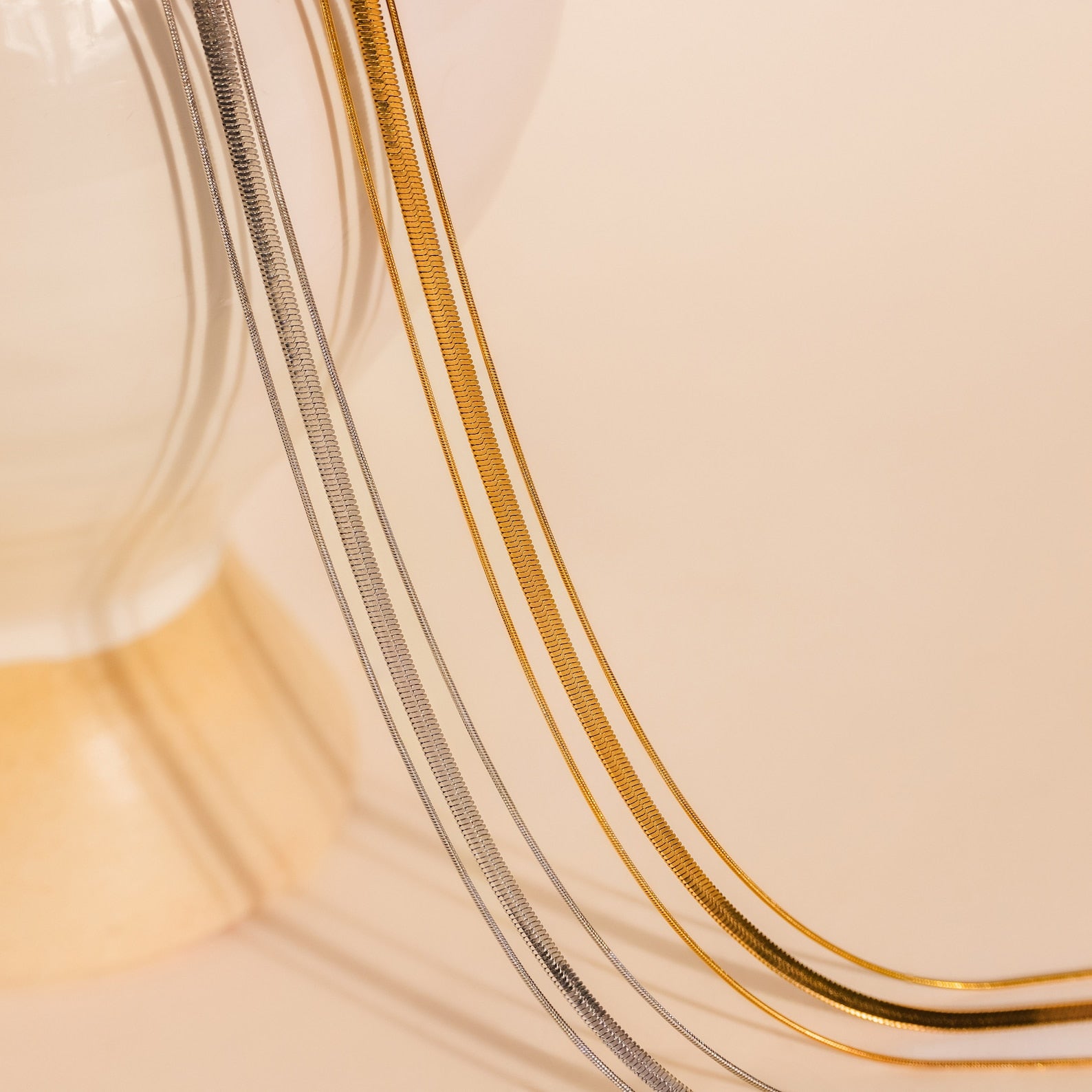 The Triple Layered Chain Necklace in gold and silver is elegantly draped over the edge of a cream-colored vase, set against a beige backdrop.