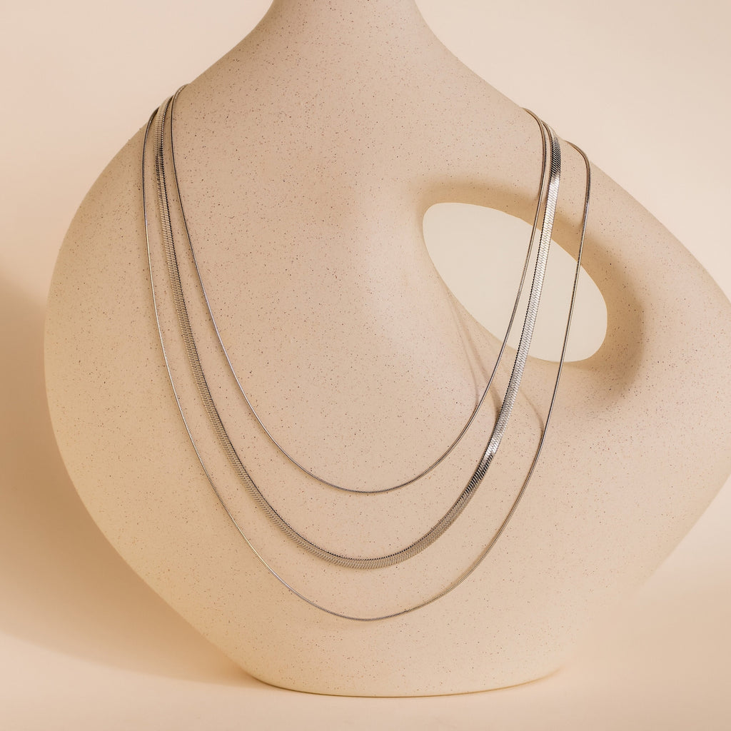 The Triple Layered Chain Necklace, featuring a sleek herringbone layer, drapes elegantly over a smooth, abstract beige ceramic vase with a large oval cutout.