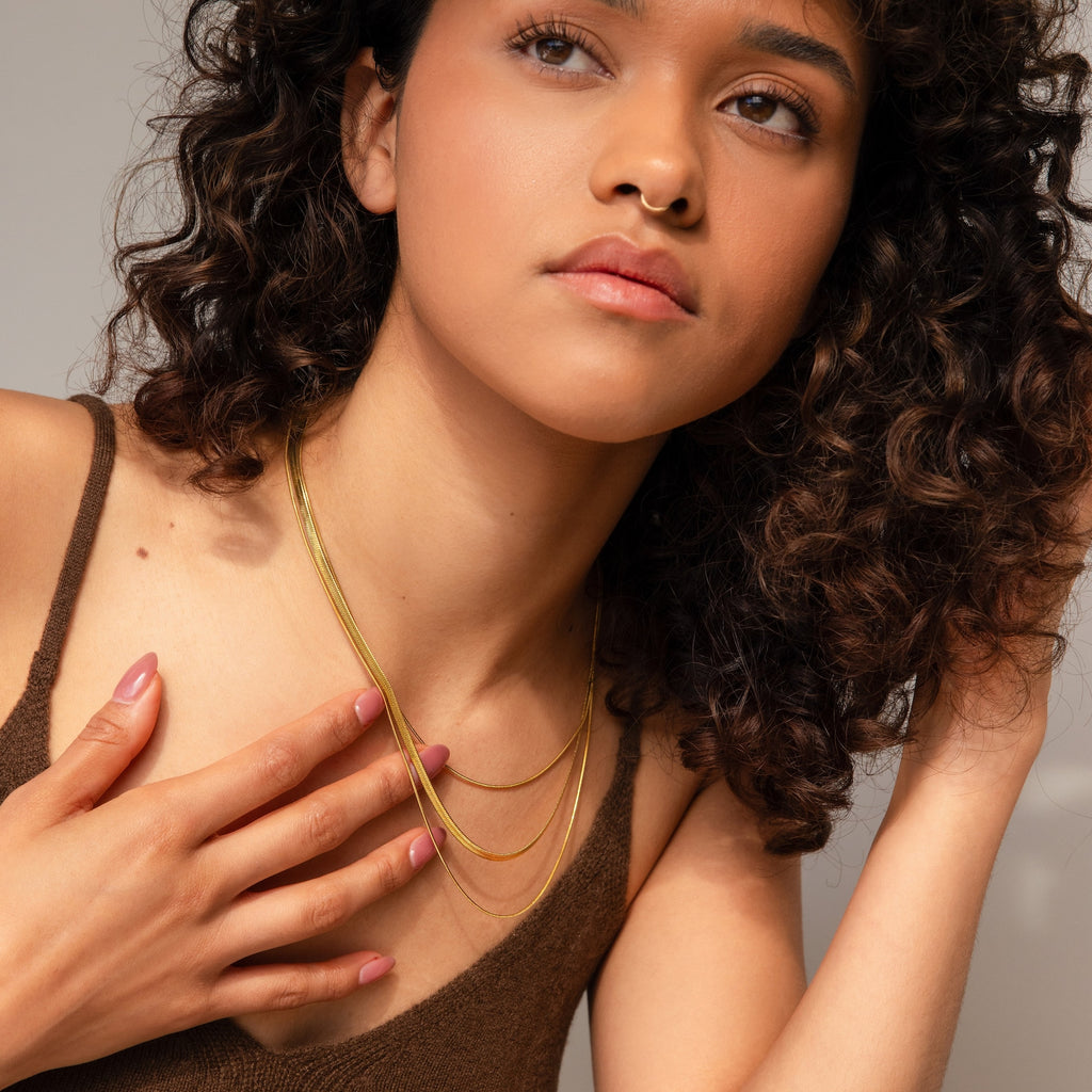 A woman with curly hair wears a brown top and the Triple Layered Chain Necklace, featuring a delicate herringbone chain, gently touching her collarbone.