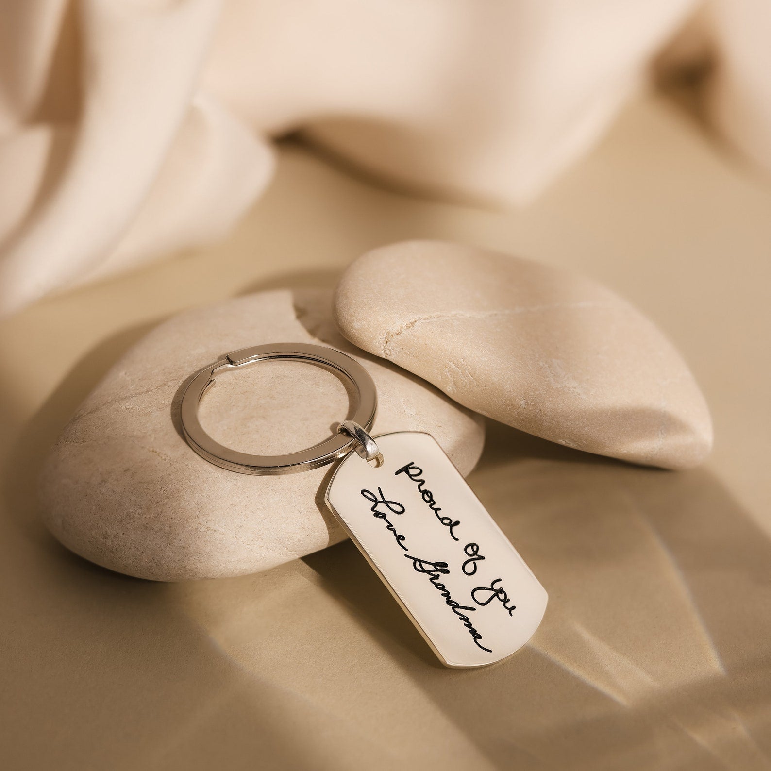 The Handwriting Dog Tag Keychain, engraved with your own handwriting, rests on beige stones and a neutral fabric—making it a perfect Father’s Day gift.