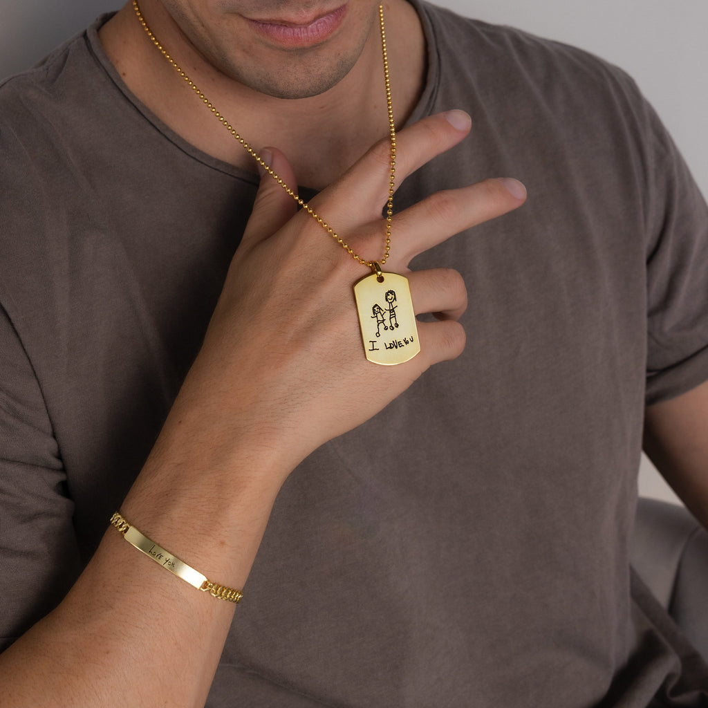 A man wearing a gold bracelet and a Men's Dog Tag Necklace holds a pendant engraved with a stick figure drawing and text—an ideal Father’s Day gift.