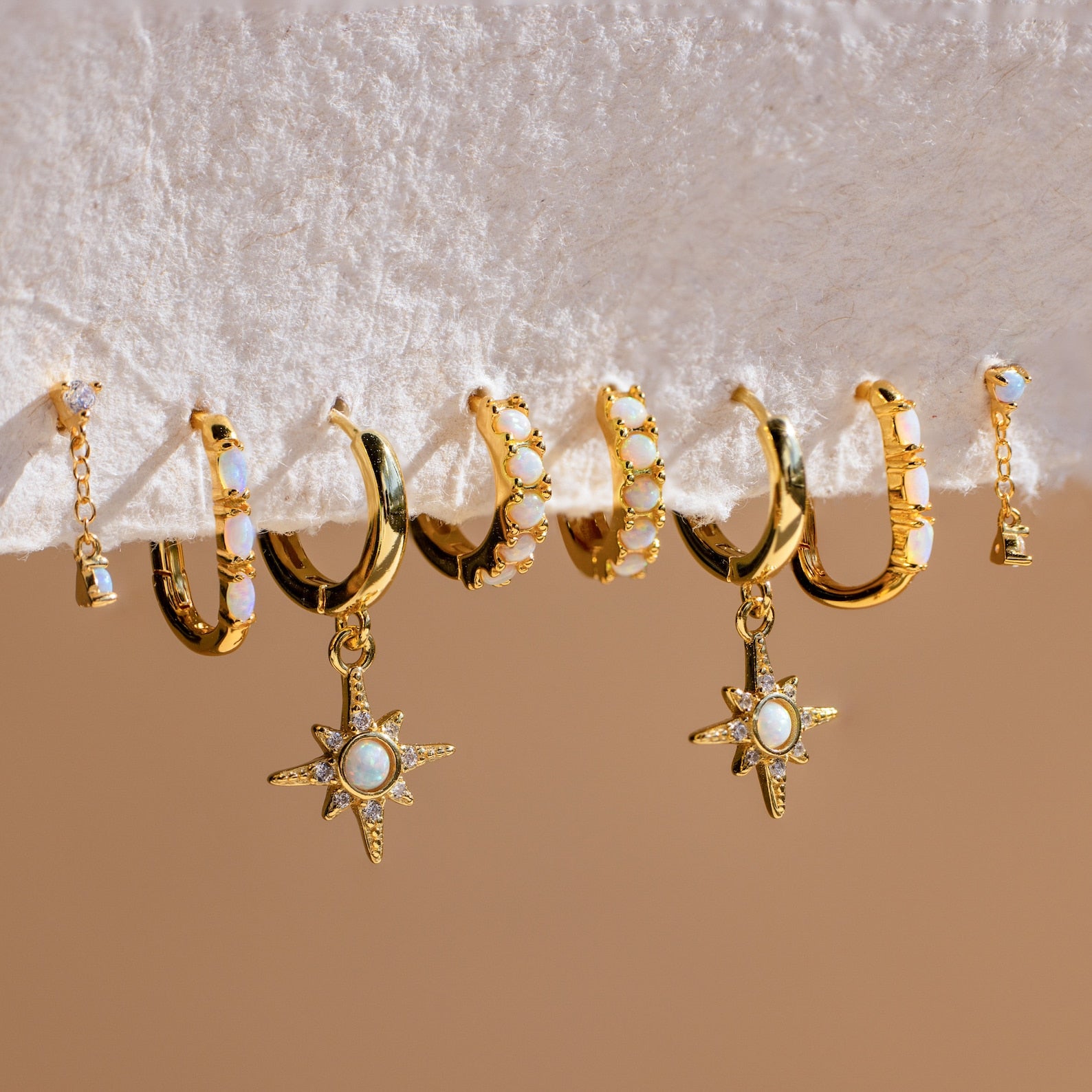 Six pairs of Dainty Opal Earrings featuring gold hoops with unique designs and gemstone accents, including genuine opal details, are displayed on a white textured surface.