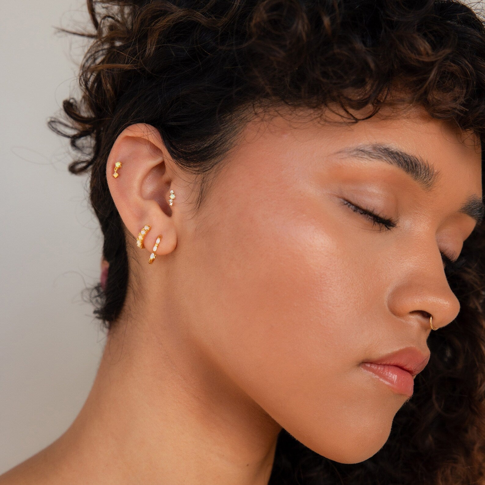 Woman with curly hair and closed eyes wearing multiple gold hoop earrings, a nose ring, and Dainty Opal Earrings.