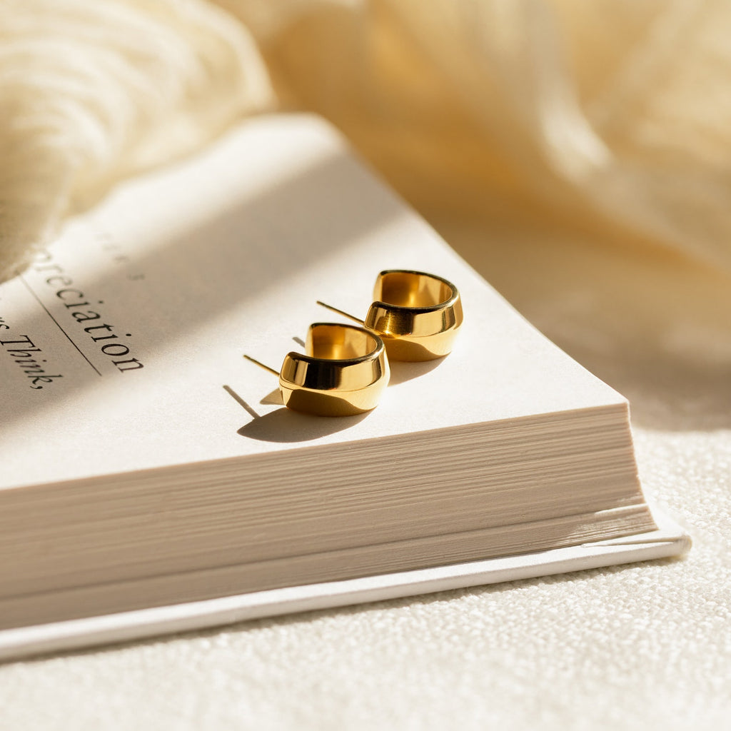 Chunky Bold Hoops shine atop an open book under soft, warm light with a blurred fabric backdrop, setting a stylish scene for statement jewelry lovers.