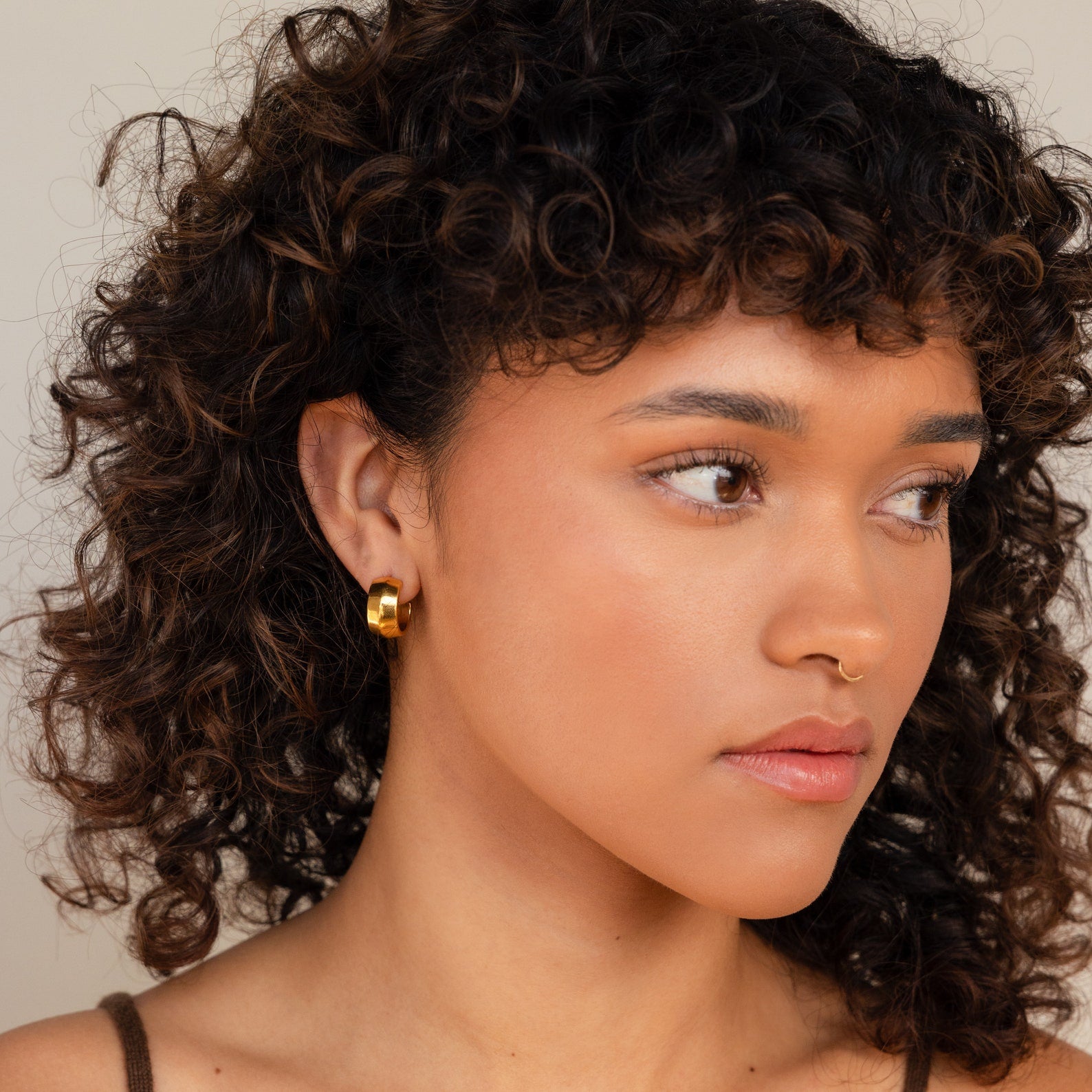A woman with curly hair wears trendy earrings—a Chunky Bold Hoop earring and a nose ring—looking slightly to the side.