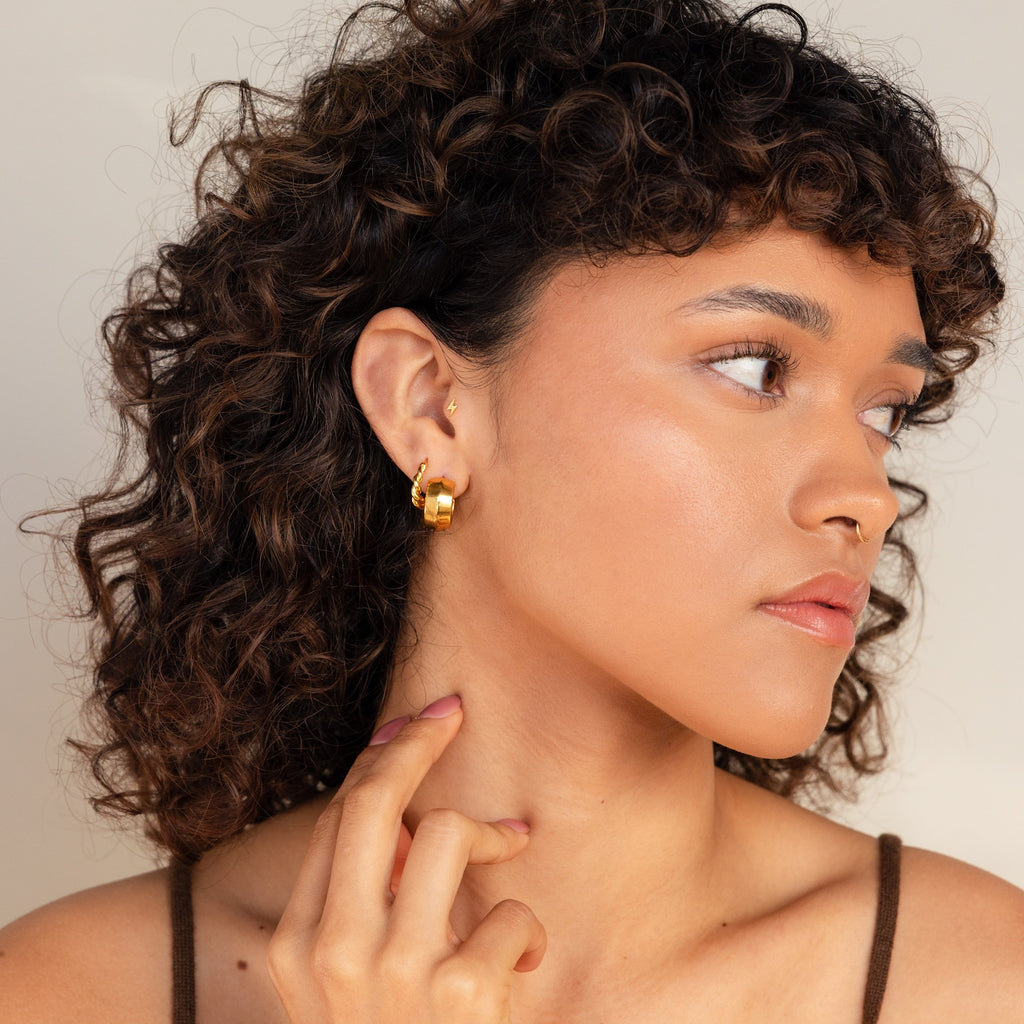A woman with curly hair wears Chunky Bold Hoops, looks to the side, and gently touches her neck.