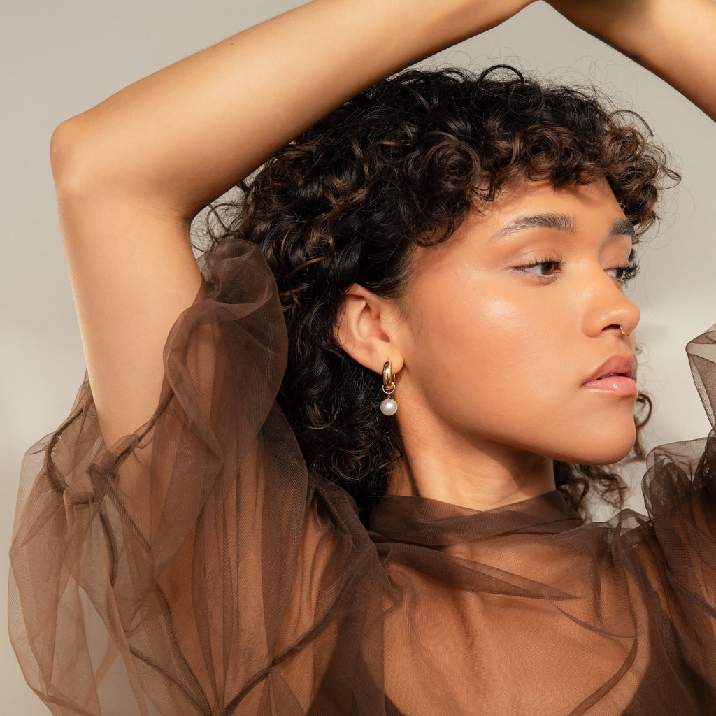 Woman with curly hair poses in a sheer brown top, wearing Bold Pearl Drop Earrings, looking to the side.