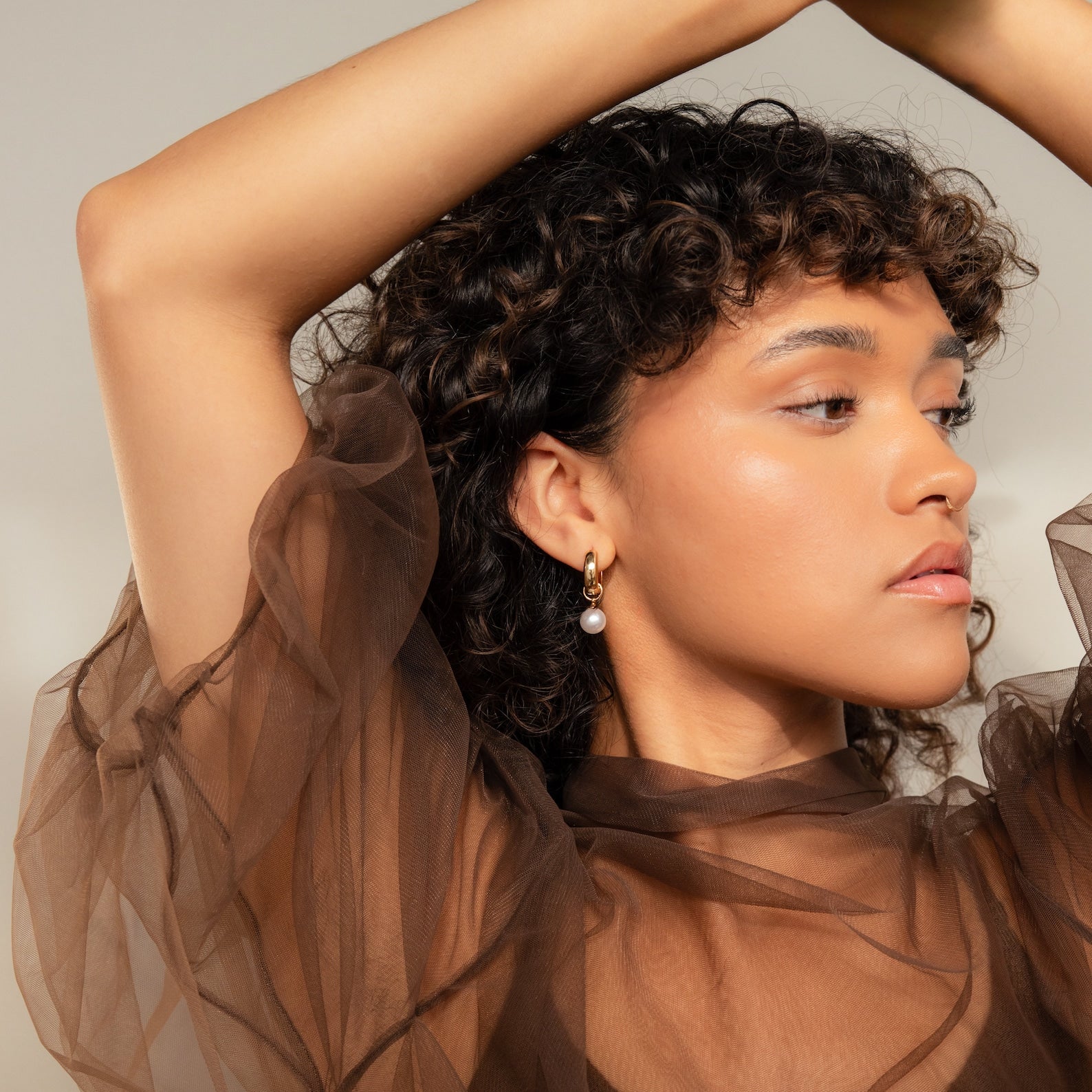 Woman with curly hair poses in a sheer brown top, wearing Bold Pearl Drop Earrings, looking to the side.