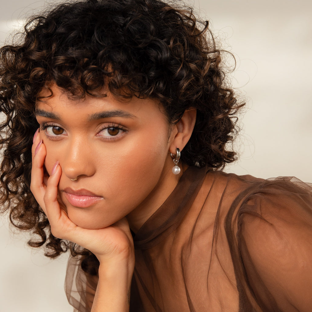 Woman with curly hair rests her face on her hand, wearing a sheer brown top and the Bold Pearl Drop Earrings.