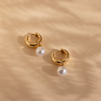 Set against a beige textured background, the Bold Pearl Drop Earrings feature gold hoops with dangling white pearls—perfect for making an elegant statement.