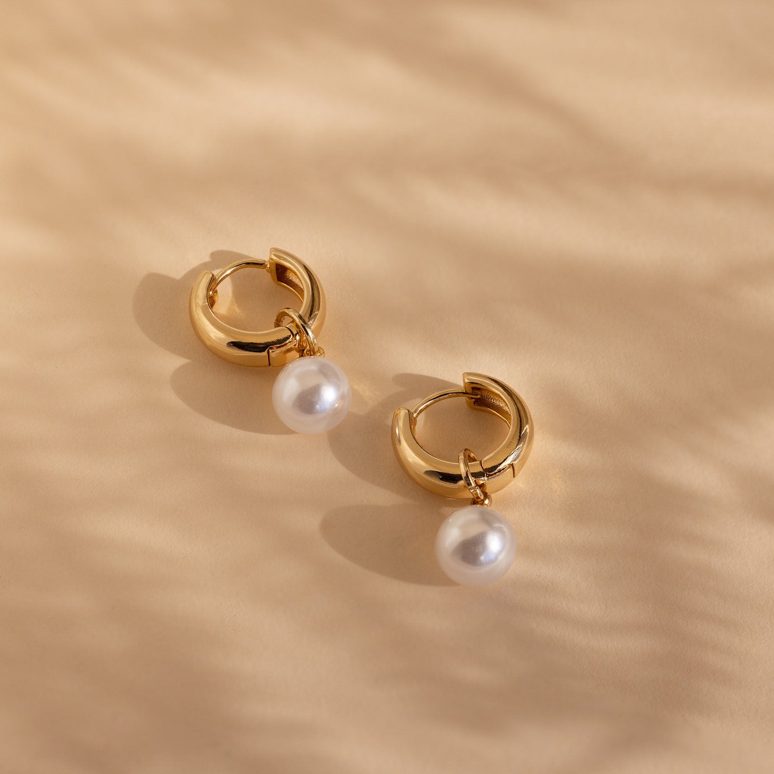 Set against a beige textured background, the Bold Pearl Drop Earrings feature gold hoops with dangling white pearls—perfect for making an elegant statement.