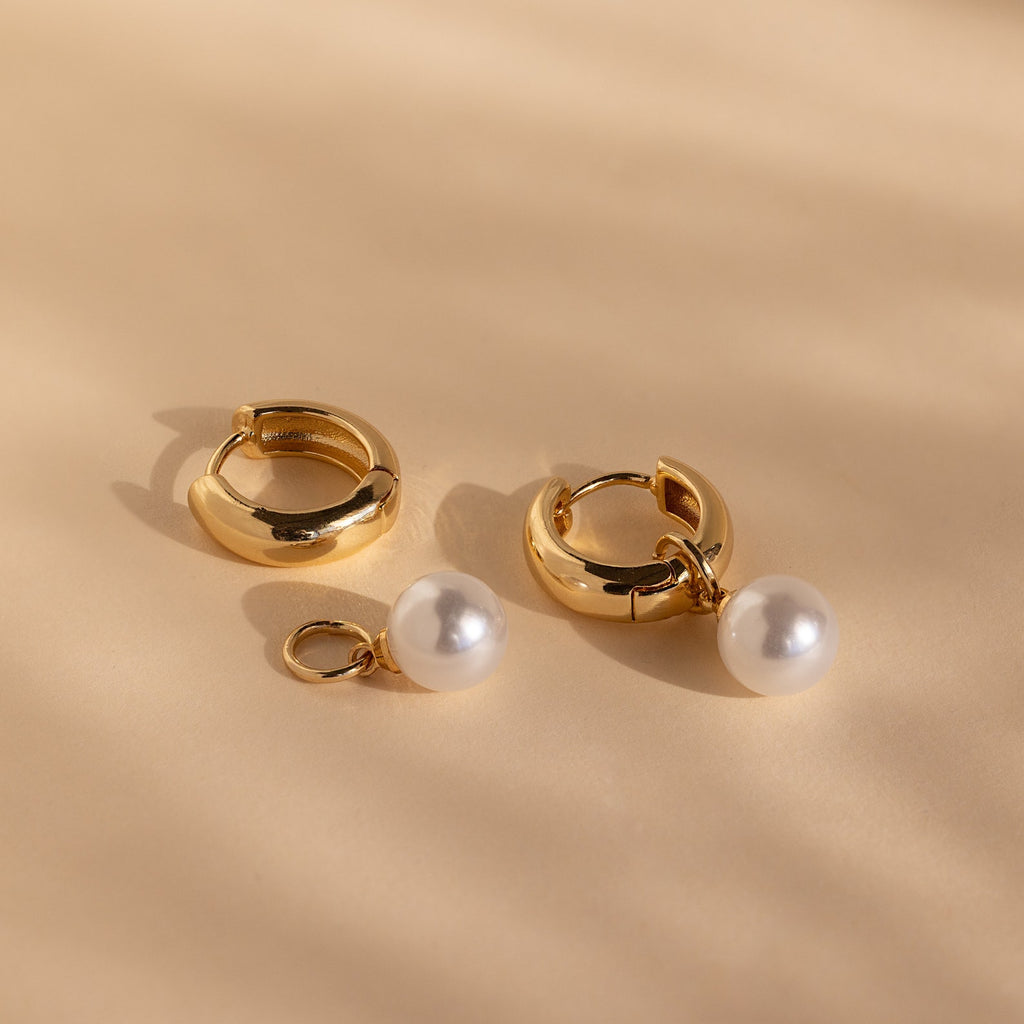 The Bold Pearl Drop Earrings feature two gold hoops, one with a detachable white pearl charm. Wear as classic gold hoops or elegant pearl drops for versatile style, shown against a beige background.