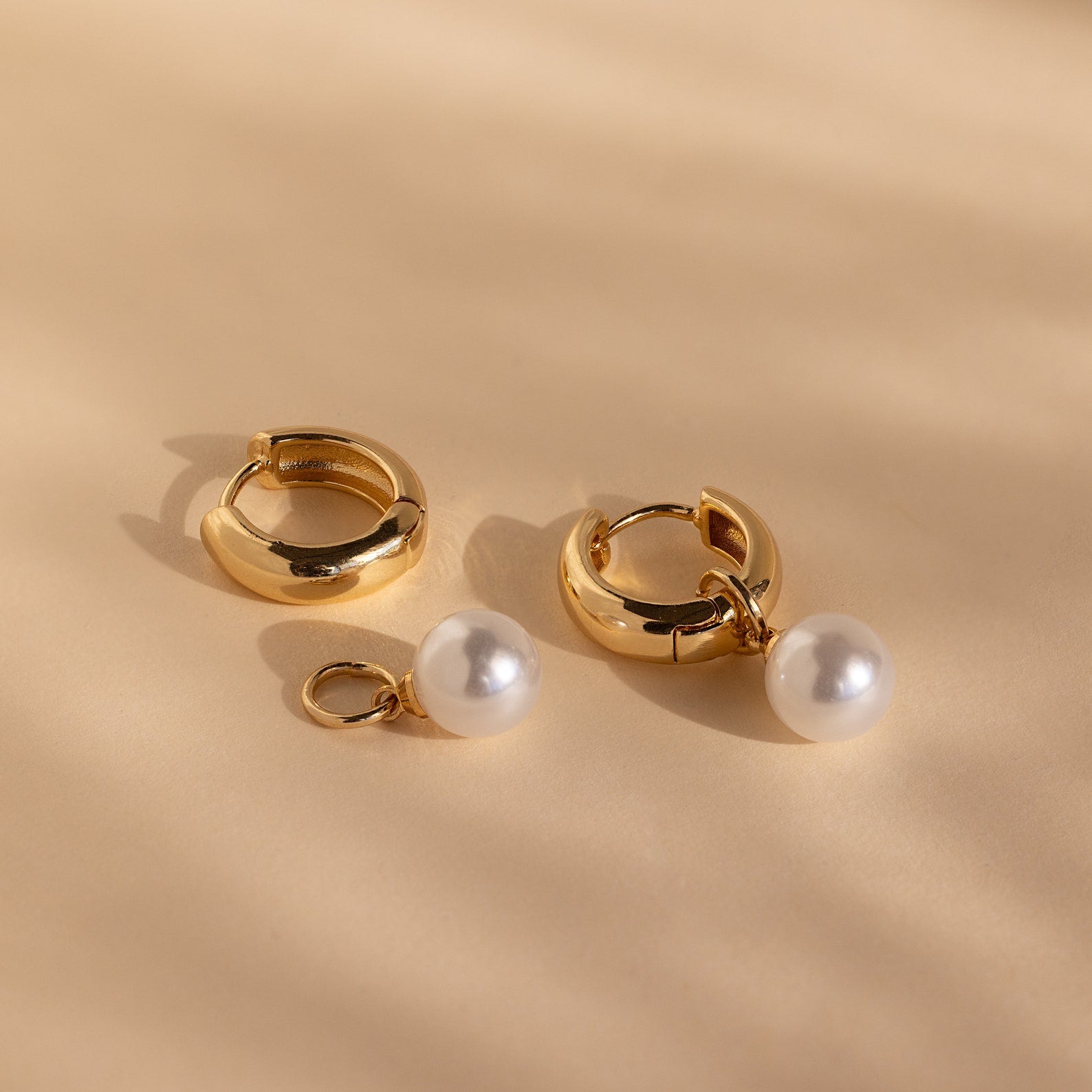 The Bold Pearl Drop Earrings feature two gold hoops, one with a detachable white pearl charm. Wear as classic gold hoops or elegant pearl drops for versatile style, shown against a beige background.