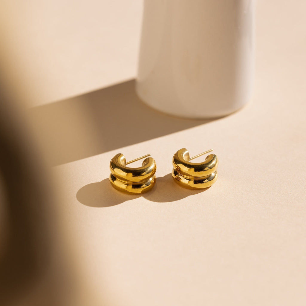 The Chunky Double Hoops rest on a light beige surface with a white vase in the background—a bold yet minimalist statement piece for any jewelry lover.