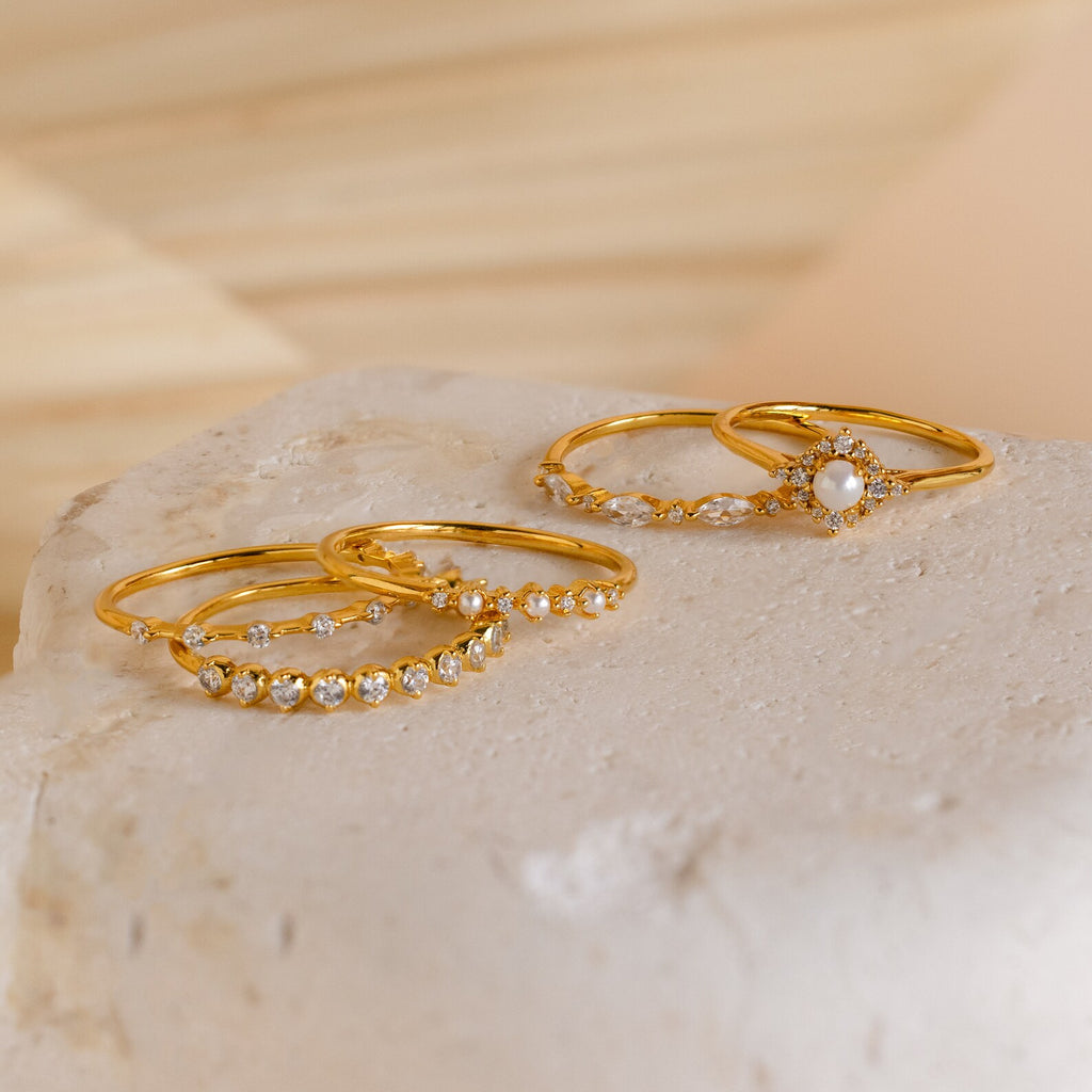 Three elegant gold rings from our Pearl Diamond Stacking Rings set, each set with small white gemstones, are showcased on a textured, light-toned stone surface.