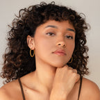 A woman with curly hair and hand on chin wears Bold Twisted Hoops, adding a bold twist design to her minimalist style.