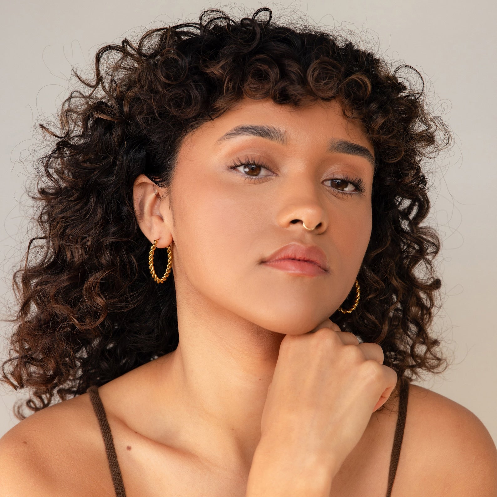 A woman with curly hair and hand on chin wears Bold Twisted Hoops, adding a bold twist design to her minimalist style.