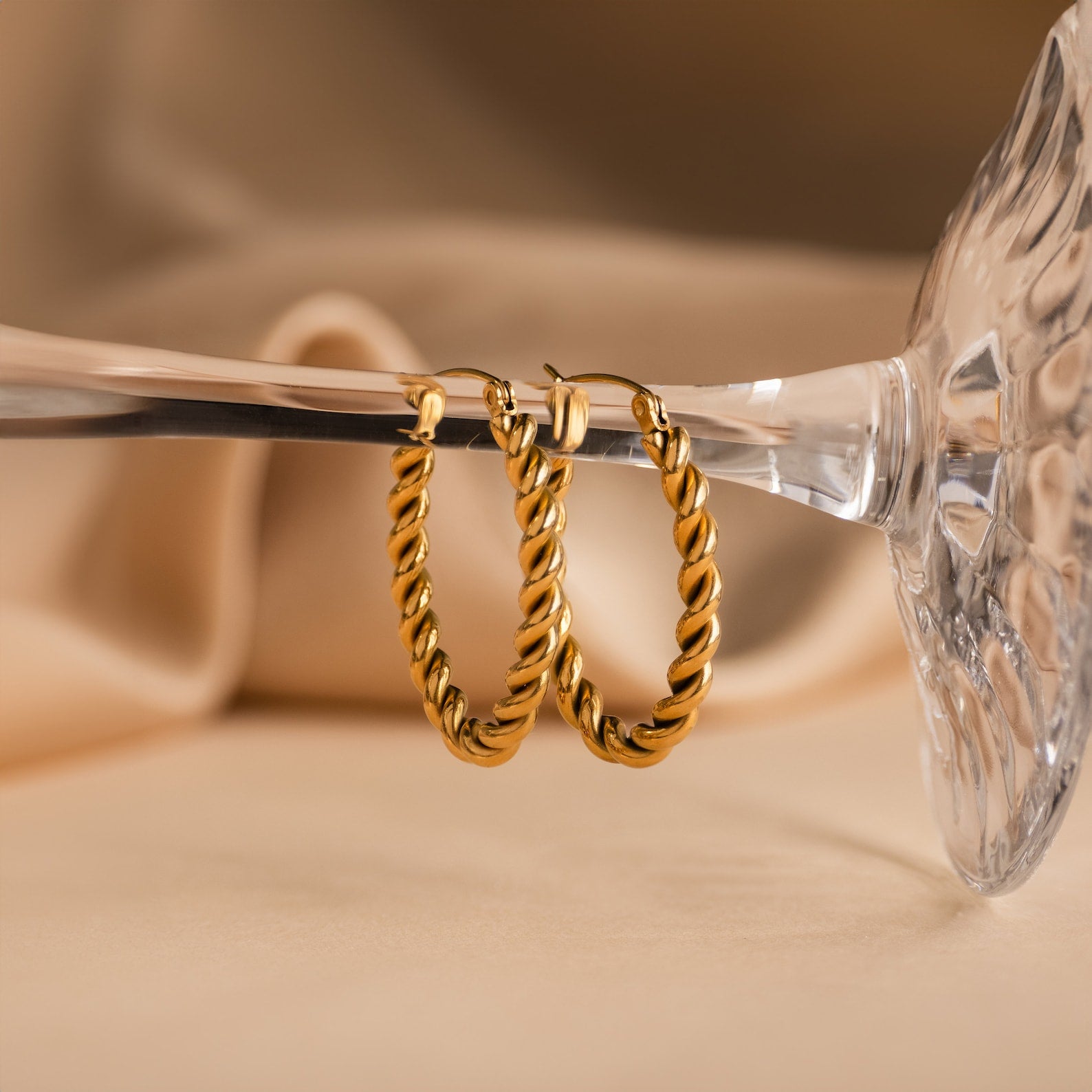 Bold Twisted Hoops, showcasing a stylish twist design, are elegantly displayed on the rim of an upside-down clear glass against a beige backdrop.