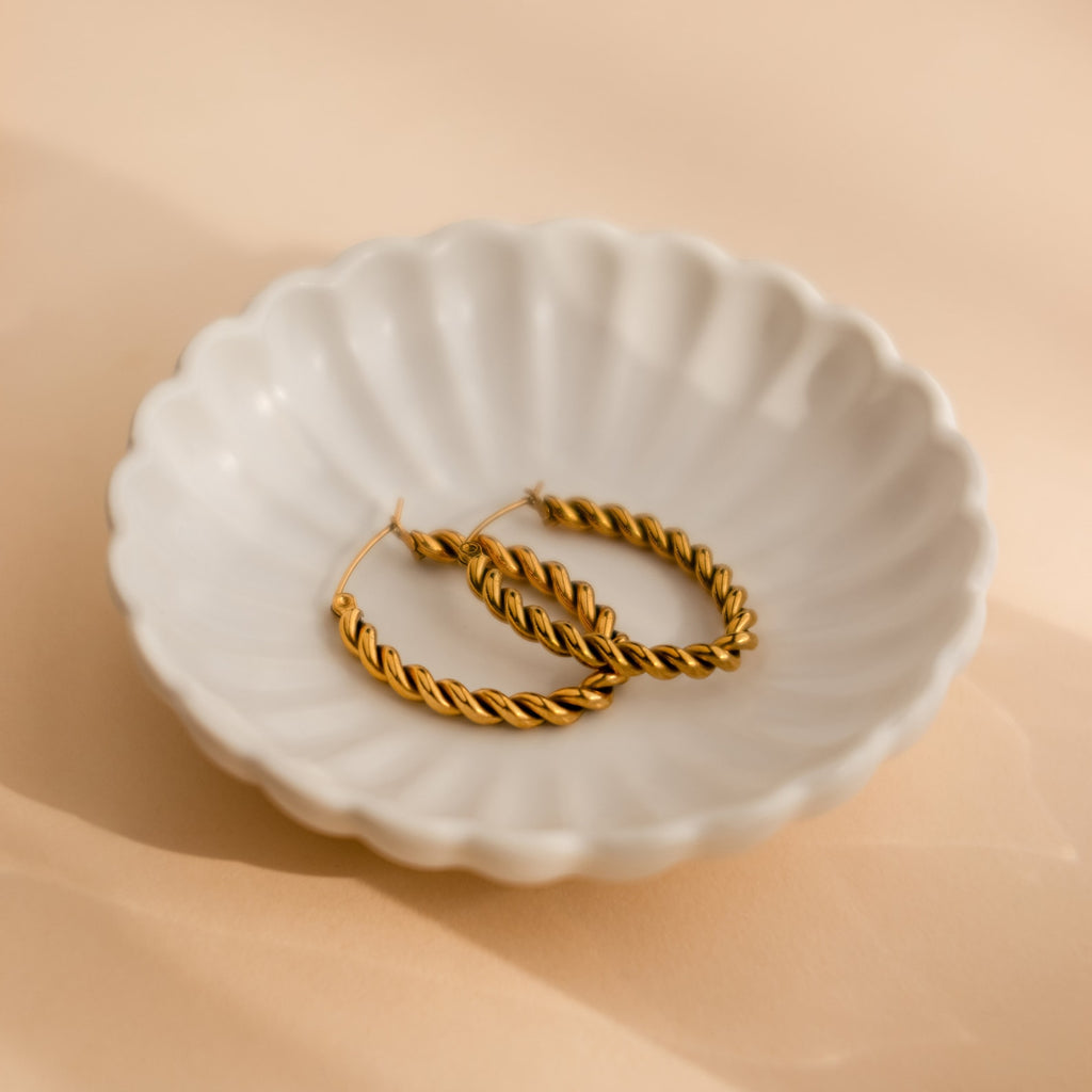 Bold Twisted Hoops, featuring a twist design, are shown on a white scalloped dish against a soft beige background—perfect minimalist earrings for any occasion.