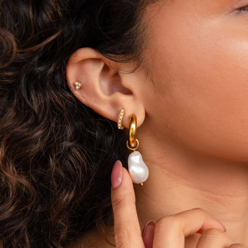 A woman with curly hair wears Baroque Pearl Drop Hoops—gold hoops featuring large baroque pearls and diamond accents—making them perfect as statement earrings for wedding jewelry.