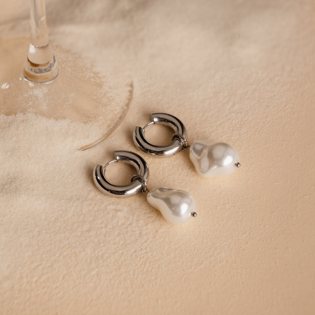 Baroque Pearl Drop Hoops with silver accents rest on soft beige sand beside a wine glass—ideal as standout earrings or elegant wedding jewelry.