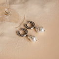Baroque Pearl Drop Hoops with silver accents rest on soft beige sand beside a wine glass—ideal as standout earrings or elegant wedding jewelry.