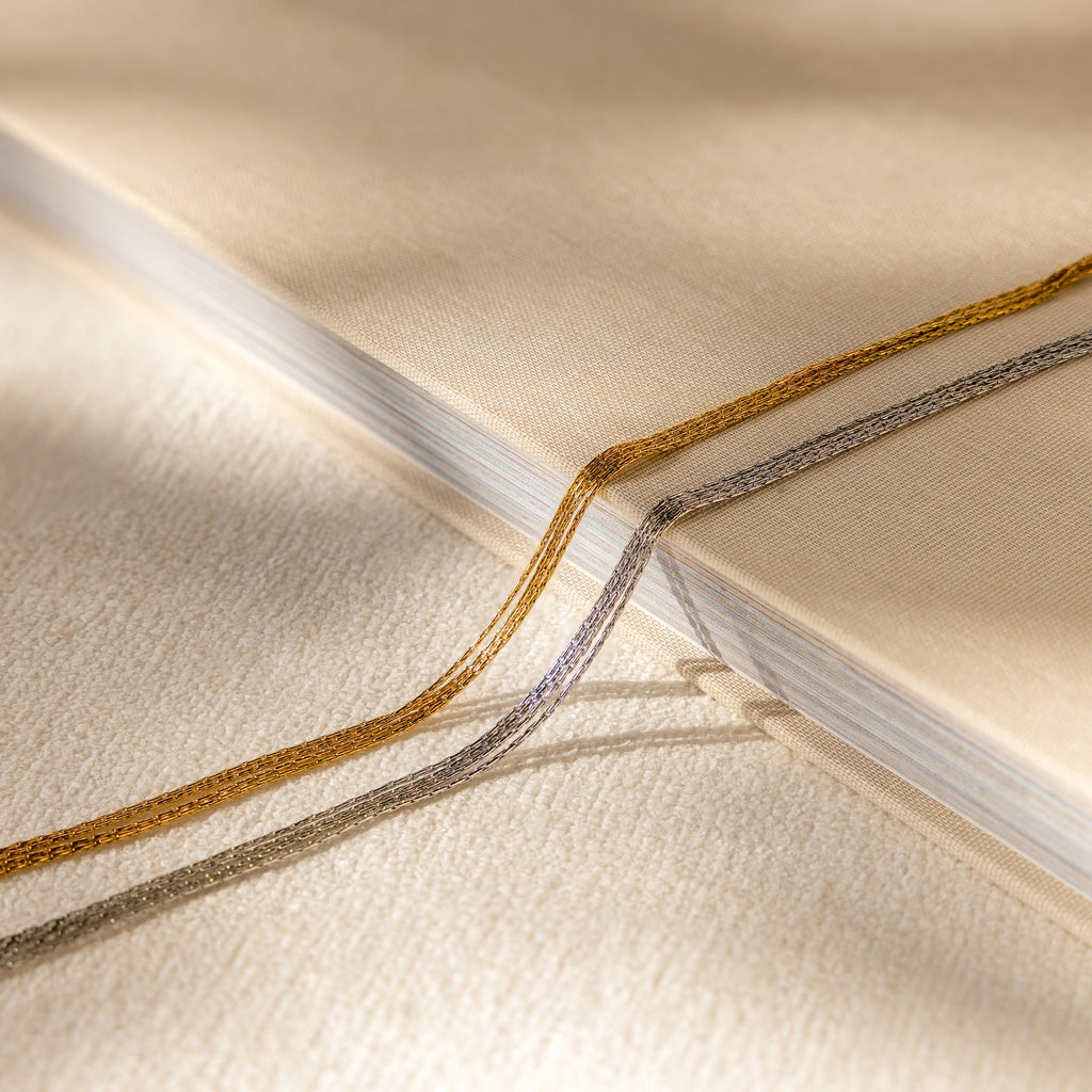 Close-up of a beige book with gold and silver ribbon bookmarks on a textured cream surface, beside a delicate Multi Strand Chain Necklace.