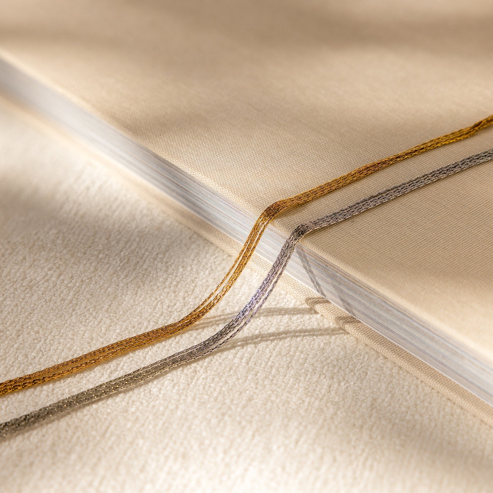 Close-up of a beige book with gold and silver ribbon bookmarks on a textured cream surface, beside a delicate Multi Strand Chain Necklace.