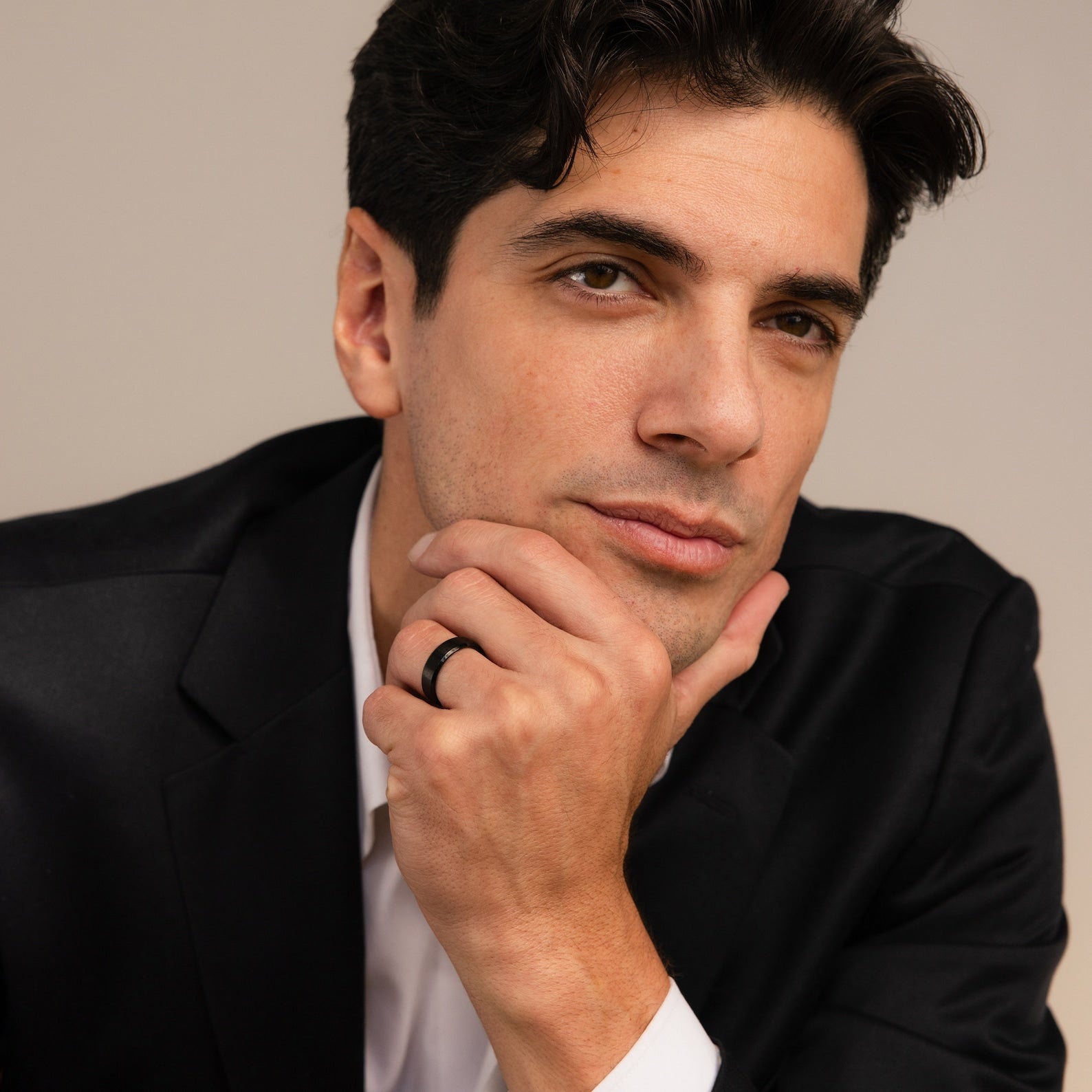 A man in a black suit and white shirt rests his chin on his hand, thoughtfully gazing at the camera while subtly showcasing the Classic Men's Black Rings Set—a sleek, minimalist touch of men’s jewelry.
