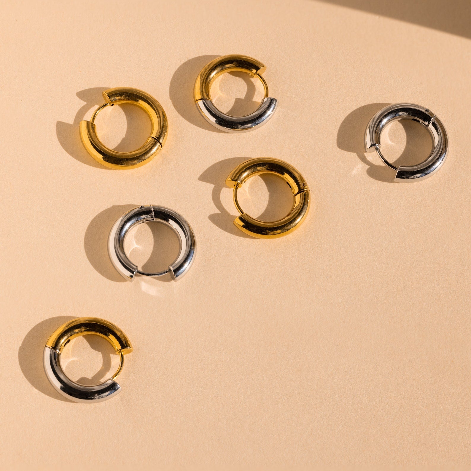 Six Chunky Hoops—three gold and three silver pairs—are displayed on a beige surface with soft shadows.